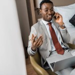 Successful handsome african american businessman with laptop talking on smartphone in hotel room