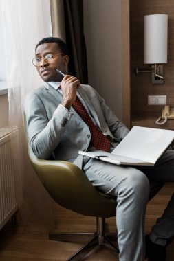 otel odasında otururken günlüğüyle yakışıklı dalgın Afrikalı-Amerikalı iş adamı
