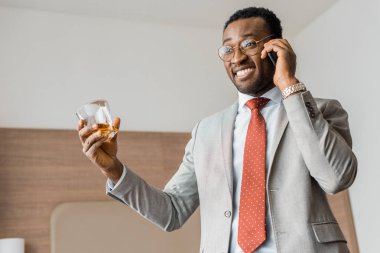 başarılı Afrikalı-Amerikalı işadamı holding bardak viski gülümseyen ve otel Smartphone cep telefonu ile konuşmak