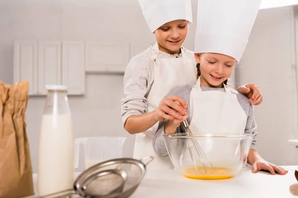 キッチンのテーブルでボウルに卵を Whisking シェフ帽子で妹を持つ少年  — 無料ストックフォト