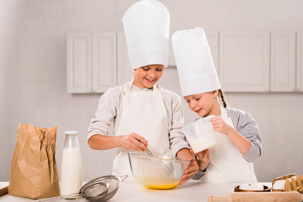 child pouring milk into bowl while brother whisking at table in kitchen 