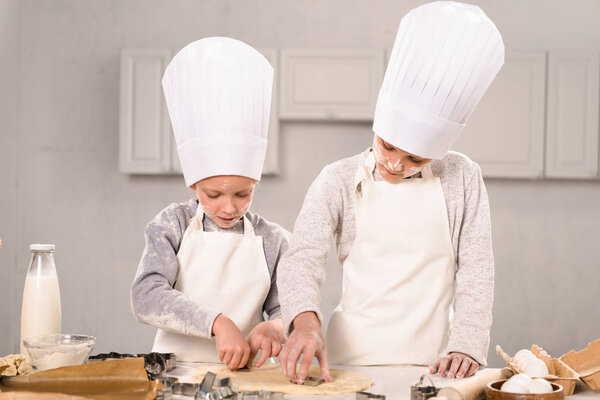 adorable little brother and sister in chef hats and aprons cutting out dough for cookies at table in kitchen