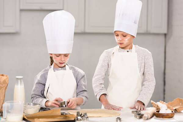 sister and brother in chef hats and aprons cutting out dough for cookies at table in kitchen