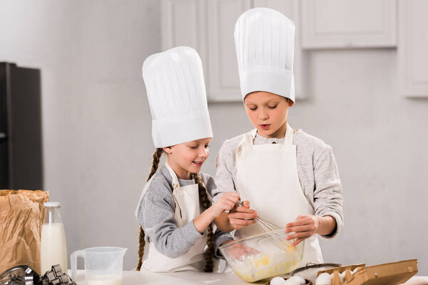 little kids in chef hats and aprons whisking eggs in bowl at table in kitchen