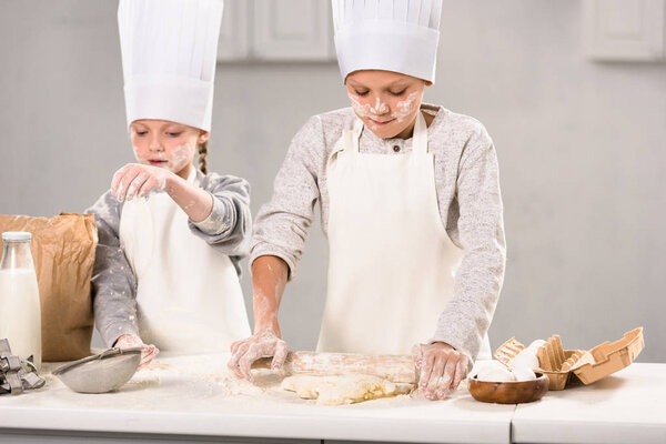 selective focus of brother and sister in aprons and chef hats making dough with rolling pin at table in kitchen
