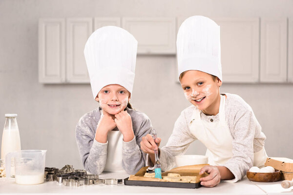 joyful children in aprons brushing cookies on baking tray in kitchen 