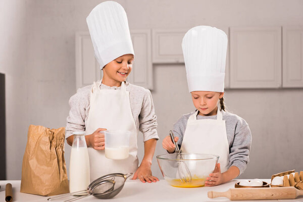 brother and sister in chef hats and aprons whisking eggs in bowl at table in kitchen