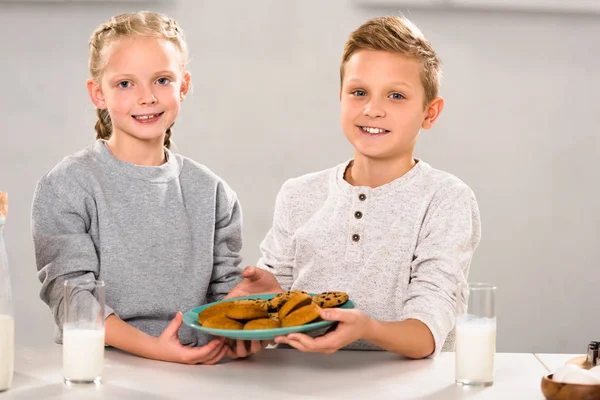 cheerful happy brother and sister holding plate with cookies near table with milk glasses