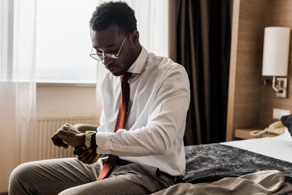 ホテルの部屋でベッドの上に座りながら腕時計を見て若いアフリカ系アメリカ人実業家  — 無料ストックフォト