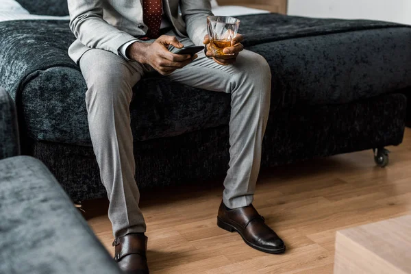 ホテルの部屋でスマート フォンを使用してウイスキーのグラスとアフリカ系アメリカ人のビジネスマンのビューをトリミング — ストック写真