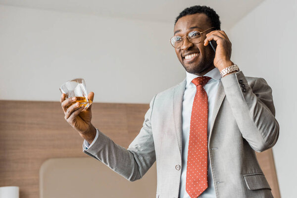 успешный улыбающийся африканский бизнесмен, держащий стакан виски и разговаривающий по смартфону в отеле
