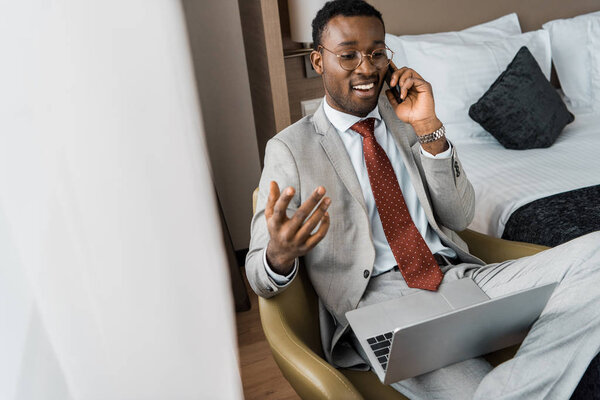 successful handsome african american businessman with laptop talking on smartphone in hotel room