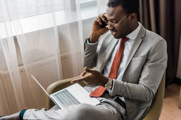 handsome smiling african american businessman talking on smartphone and pointing at laptop in hotel room