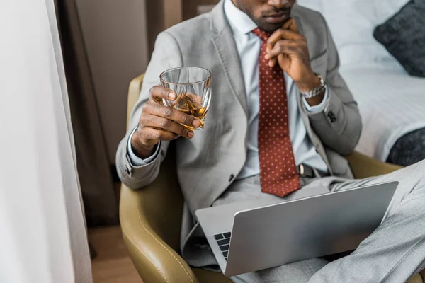 裁剪视图沉思的非洲裔美国商人与一杯威士忌看着笔记本电脑在酒店房间 — 图库照片