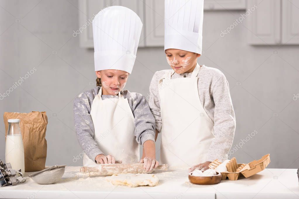 sister and brother in aprons and chef hats making dough with rolling pin at table in kitchen