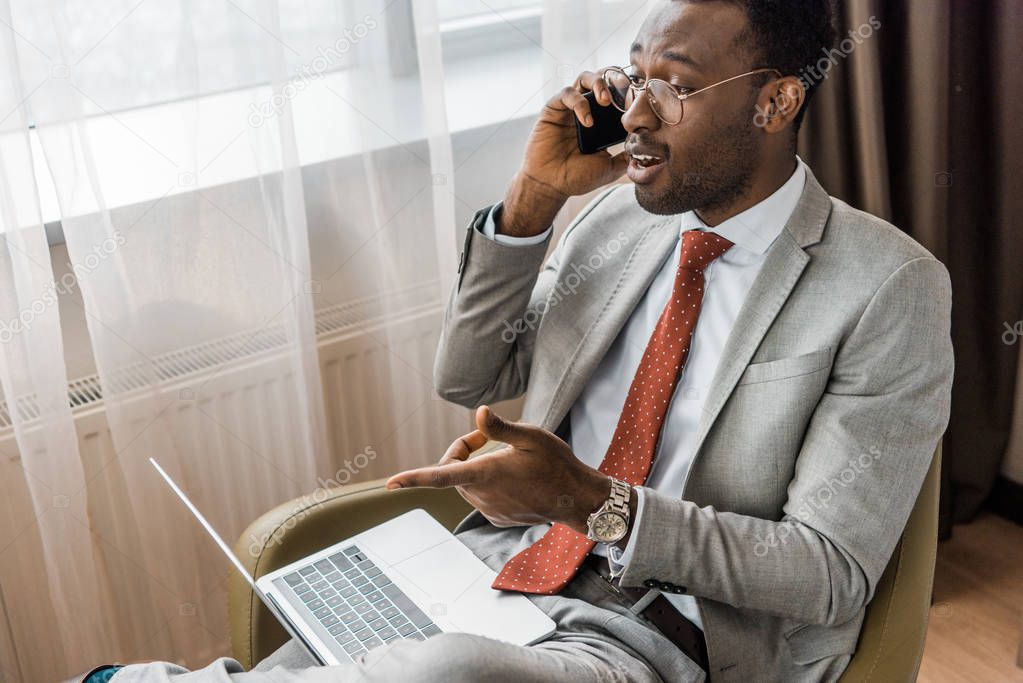 successful african american businessman talking on smartphone and pointing at laptop 