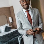 Glimlachen van Afro-Amerikaanse zakenman gebruikend smartphone in hotelkamer