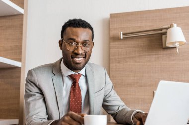 smiling african american businessman with laptop on coffee break in hotel room clipart