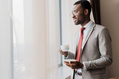 otel odasında pencere yakınındaki kahve tutan takım elbiseli yakışıklı işadamı