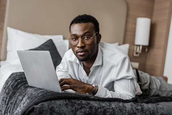 ホテルのベッドの上のラップトップで働く眼鏡のハンサムなアフリカ系アメリカ人ビジネスマン — ストック写真