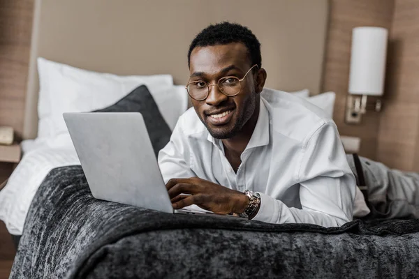 ホテルのベッドの上のラップトップで働く眼鏡のアフリカ系アメリカ人のビジネスマンを笑顔 — ストック写真
