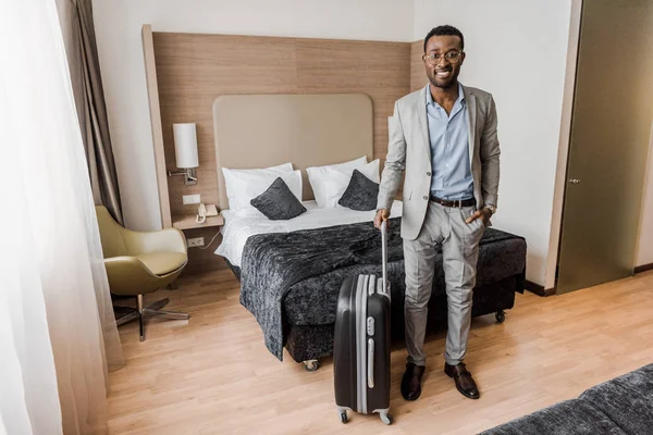 ベッドとホテルの部屋でスーツケースを持ったエレガントなアフリカ系アメリカ人ビジネスマン — ストック写真