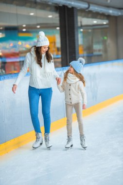 smiling mother and daughter holding hands and skating on ice rink together clipart