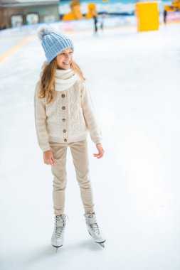 cheerful kid in sweater and skates looking away on skating rink clipart