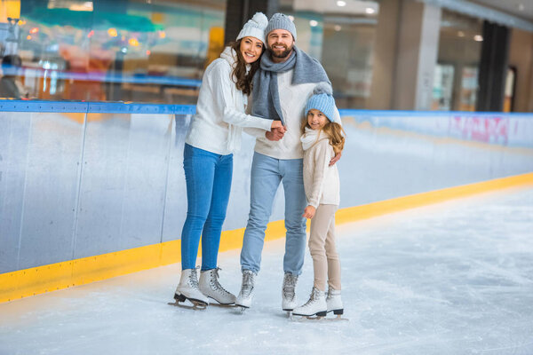 smiling parents and daughter in sweaters looking at camera on skating rink