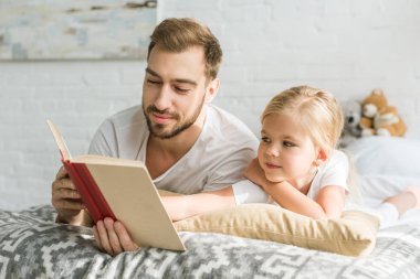 mutlu baba ve sevimli küçük kız yatakta yatarken ve kitap okuma 
