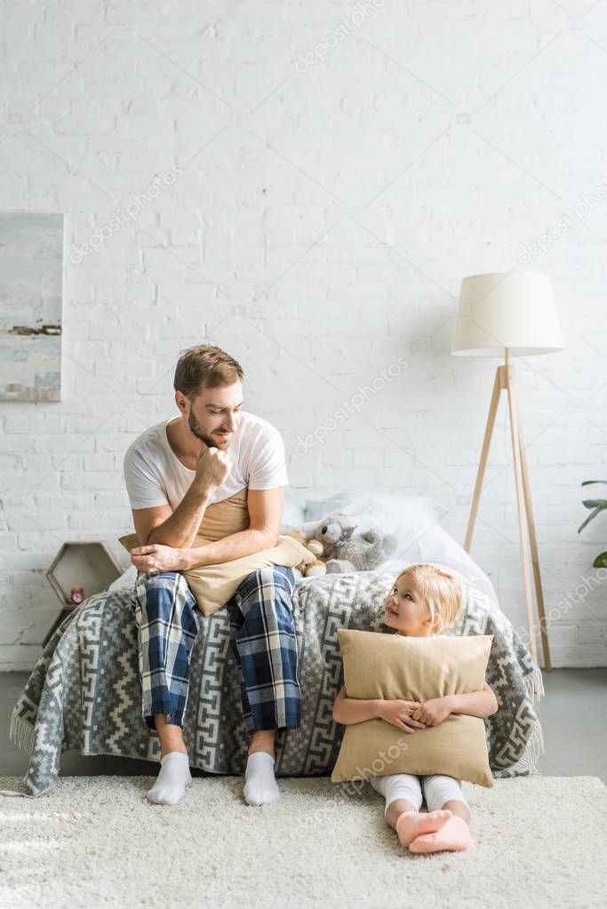 father and daughter holding pillows and smiling each other while sitting in bedroom