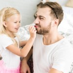 Мила маленька дочка застосовує червону помаду до щасливого бородатого батька вдома