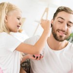 Roztomilá malá dcera použití make-up, šťastný vousatý otec doma