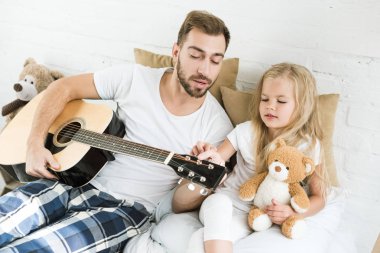 Baba ve sevimli küçük kız ile oyuncak ayı akustik gitar yatağa arıyorsunuz 