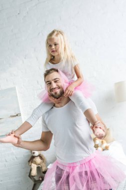 Kameraya gülümseyen ve pembe tutu etek boyunda sevimli küçük kızını taşıyan mutlu baba 