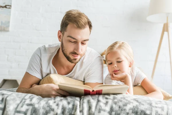 父と愛らしい小さな娘のベッドに横になっていると 本を読んで — ストック写真