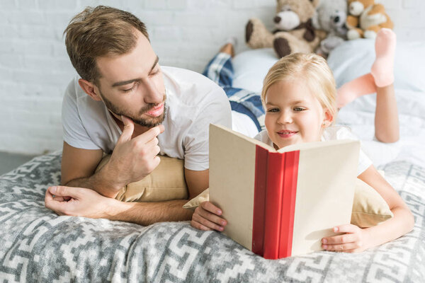 father looking at cute little daughter reading book on bed