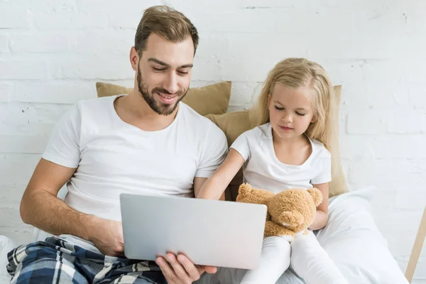 快乐的父亲和可爱的小女儿与泰迪熊一起使用笔记本电脑在卧室 — 免费的图库照片