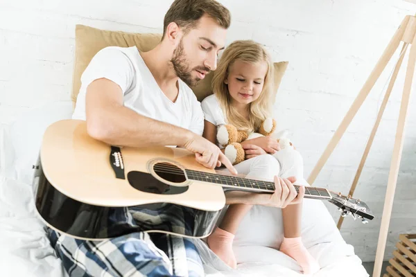 father pointing at acoustic guitar to smiling little daughter with teddy bear on bed