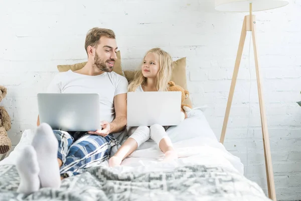 幸福的父亲和女儿微笑对方 而使用笔记本电脑在床上 — 图库照片
