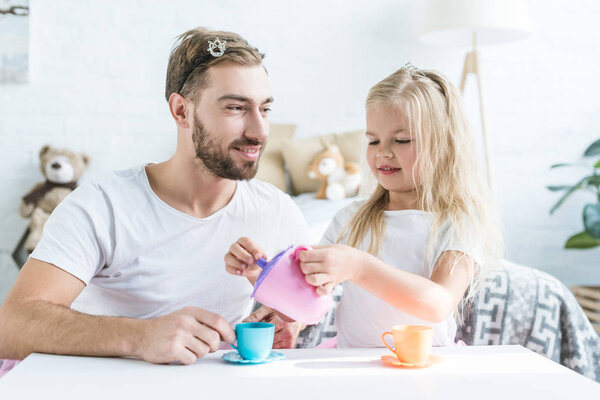 happy father looking at smiling little daughter pouring tea from toy kettle