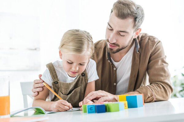 father looking at cute little daughter writing in workbook and learning at home