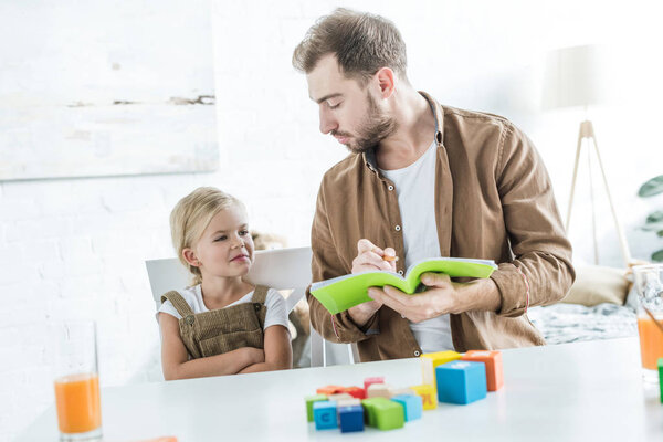 father writing in workbook and looking at little daughter learning math with colorful cubes