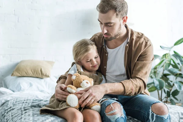 父亲看着可爱的小女儿坐在床上与泰迪熊 — 图库照片