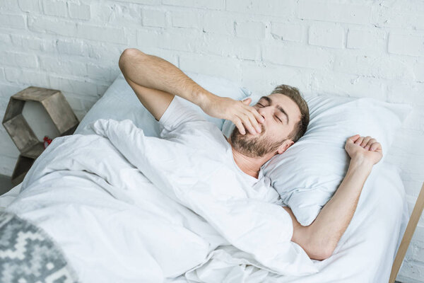 handsome young man yawning and stretching while lying in bed 