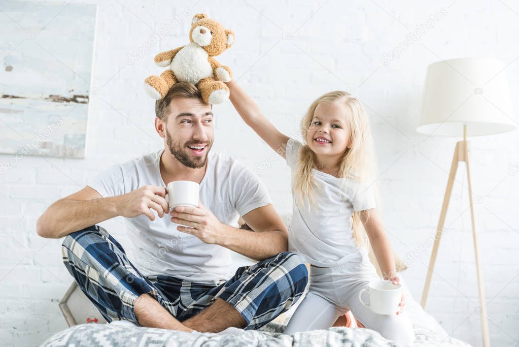 cute little daughter playing with teddy bear and smiling at camera while happy father drinking tea on bed