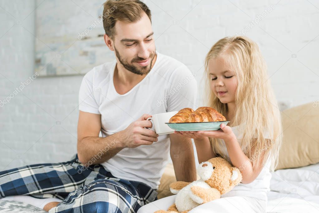 happy father holding cup of coffee and looking at little daughter with tasty croissants on plate  