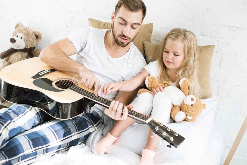 high angle view of father and cute little daughter with teddy bear looking at acoustic guitar on bed 