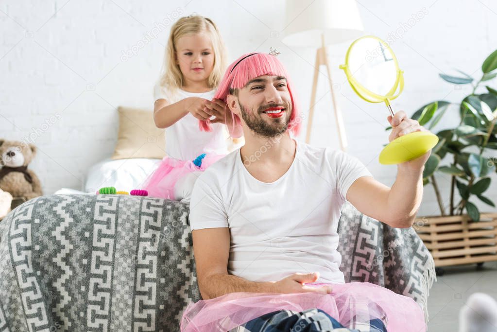 adorable little daughter in tutu skirt playing with happy father in pink wig looking at mirror     