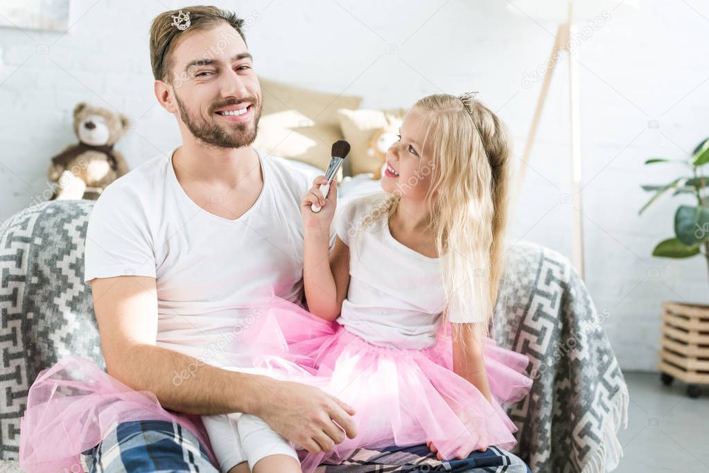 adorable little daughter in pink tutu skirt applying makeup to happy father 